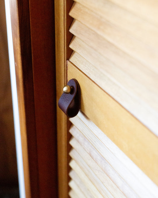 Brown leather cabinet pull with a brass mounting ball on a louvered wood door