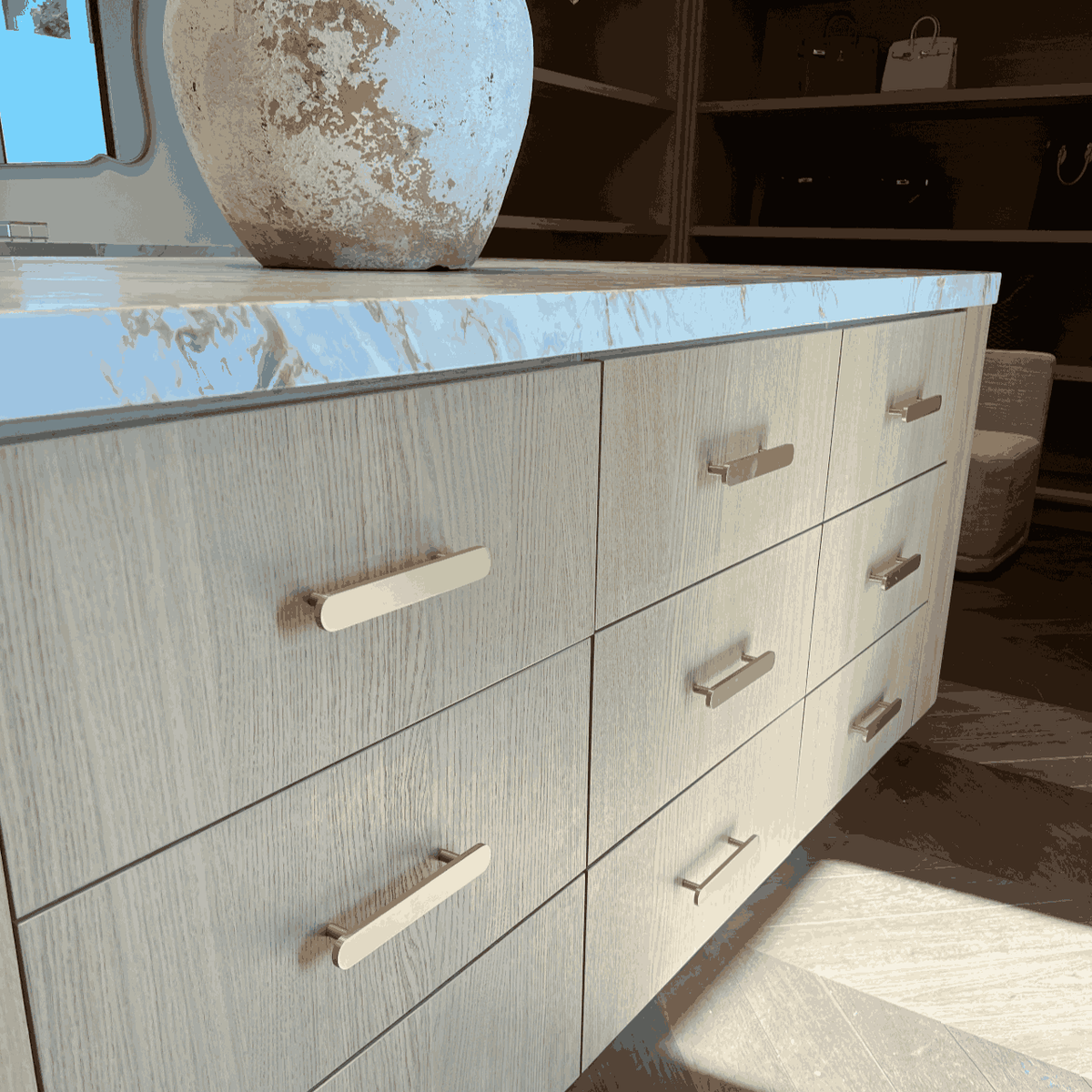 Straight brass-colored pulls on pale wood kitchen drawers beneath a veined countertop.